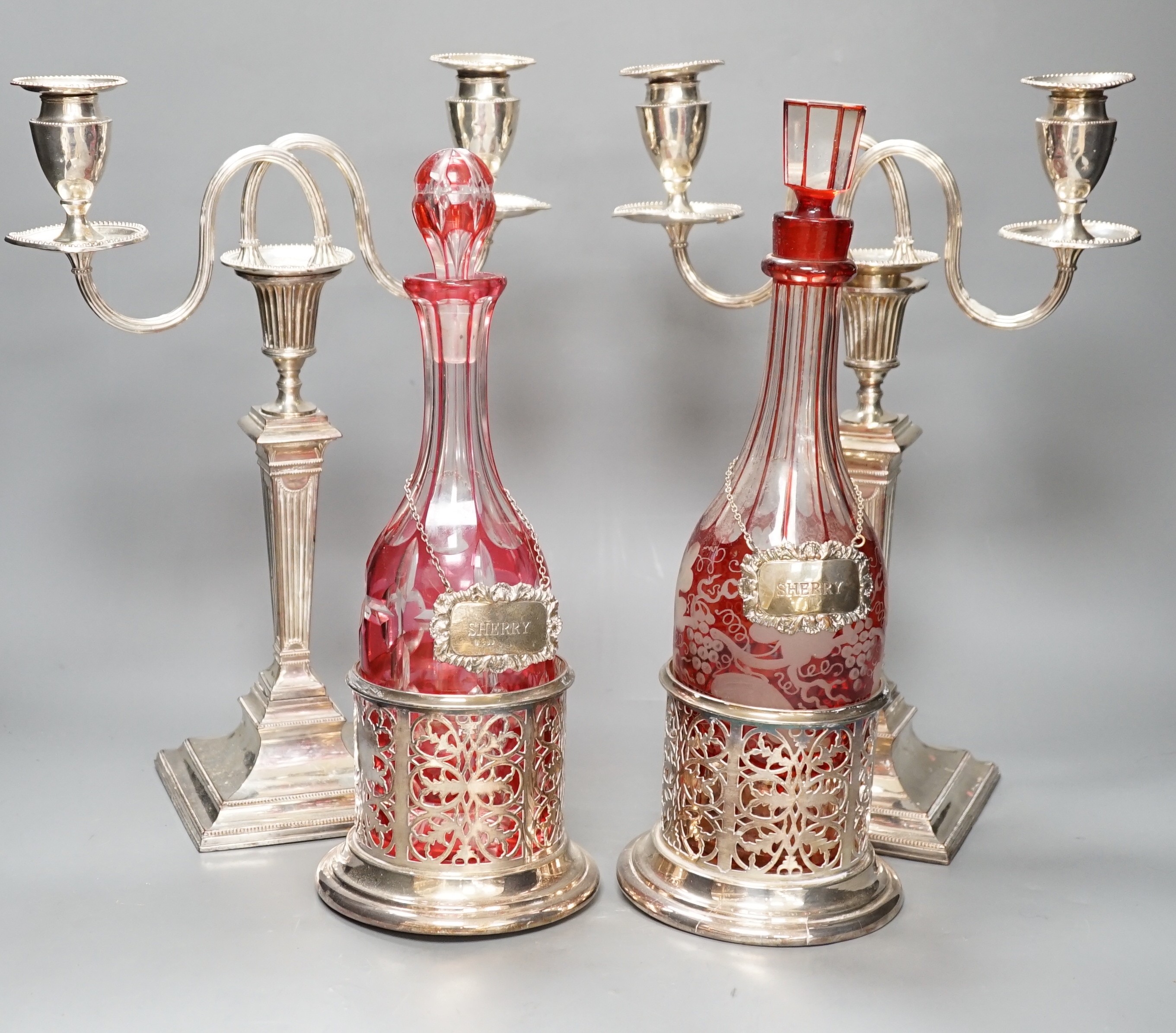 A pair of silver plated two branch, two light candelabra and a pair of silver plated decanter coasters with two ruby flash glass decanters with plated wine labels.